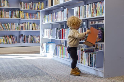 A toddler with curly hair wearing a sweater and boots is selecting a book from a shelf in a library. Cleveland, Ohio, USA