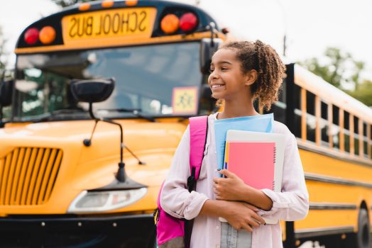 Student preparing to go to school after summer holidays holding books and notebooks standing next to the school bus.