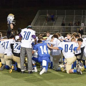 Football coach Joe Kennedy standing, holding up helmets and praying over a large group of kneeling players on the football field