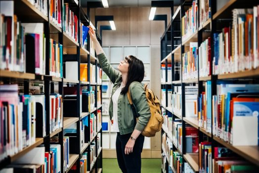 A young student reaching to select a book from the top shelf in a public library.