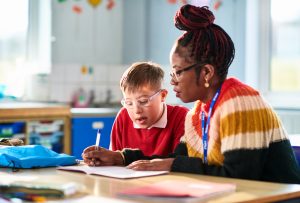 Female support worker assisting child with special educational needs, writing in his book as he watches and concentrates