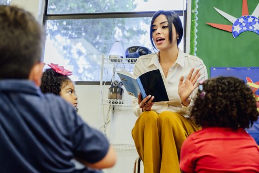 Hispanic schoolteacher reading aloud to her young students Young female Hispanic schoolteacher reading aloud to elementary aged students sitting around her on the floor.